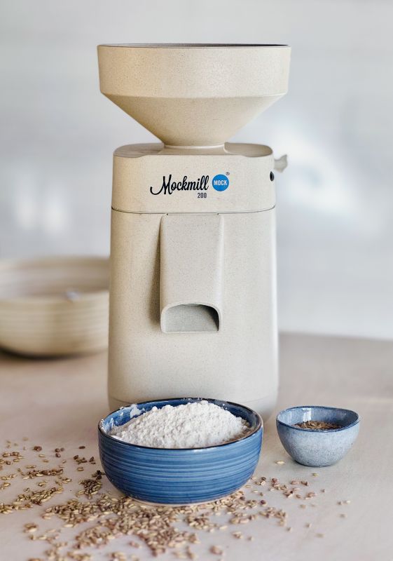Mockmill 200 stone grain mill on a kitchen counter with a blue ceramic bowl of freshly milled white flour and scattered grain kernels